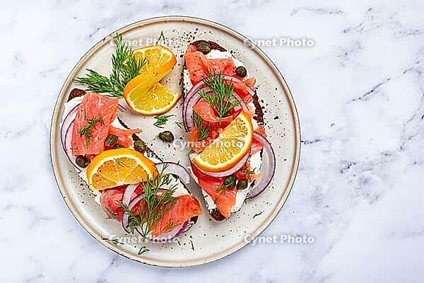 Sandwich with freshly smoked salmon, on black bread, with cottage cheese, with herbs and red onions, capers and pieces of citrus fruits, on a marble countertop, breakfast, no people [IBR124585362]