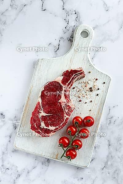 Raw rib eye steak, cowboy steak, on a white wooden chopping board, marble table, top view, no people [IBR124585364]