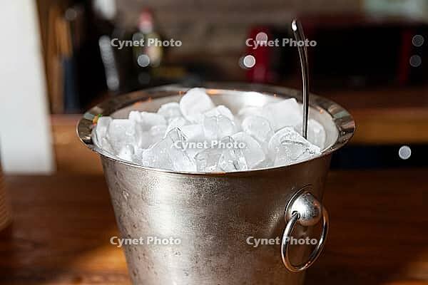 A metal bucket filled with ice cubes sits on a wooden countertop, with a kitchen background. perfect for refreshing drinks or parties, it adds a touch of chill to indoor gatherings [IBR124626866]