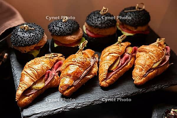 A selection of gourmet mini sandwiches featuring black sesame seed buns and croissants filled with deli meats and pickles. ideal for elegant catering events [IBR124626875]