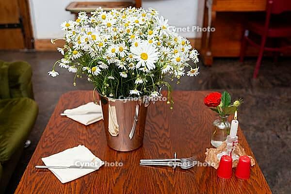 A wooden table elegantly set with a centerpiece of fresh daisies in a metallic vase. the table also features a single red rose, a lit candle, and neatly folded napkins, creating a cozy and inviting atmosphere [IBR124626899]