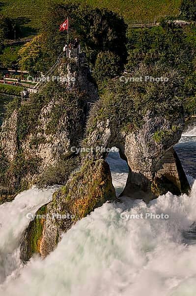 Rhine Falls in Neuhausen with rock formation in the middle of the waterfall, rock hill with hole above the water vortex, Swiss plume at the top of the rock formation with visitor platform, masses of water flow whirled up and foaming around the rock, green [IBR124636284]
