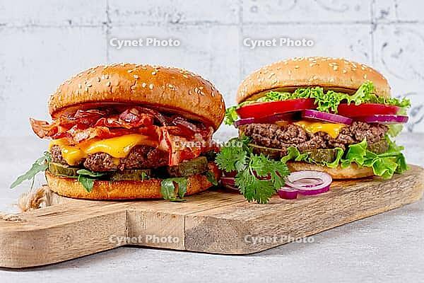 Two burgers with beef patty, fried bacon, cheddar cheese, tomatoes, on a light table [IBR124748953]