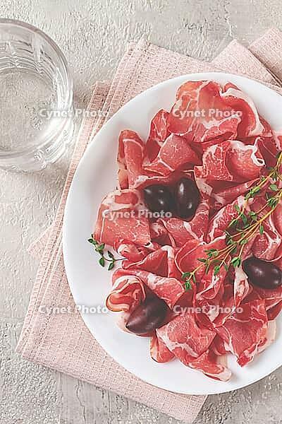 Freshly cut dried meat, served with olives and herbs, on a white plate, light background, no people [IBR124748955]