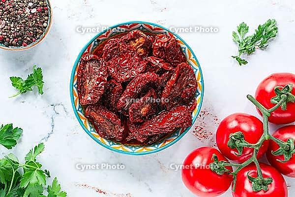 Dried tomatoes, in a ceramic bowl, with fresh herbs and spices, on a white marble surface, top view, no people [IBR124748958]
