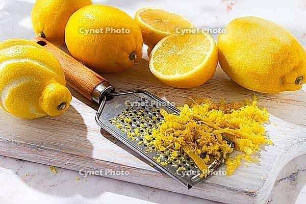 Lemons are placed on a wooden cutting board. Some lemons are whole while others are cut into slices. A zester is used to create lemon zest. The scene shows a bright kitchen [IBR124748966]