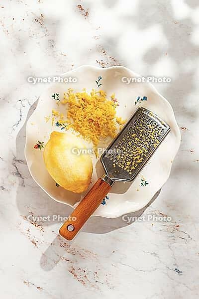 A lemon is on a small decorative plate next to finely grated lemon zest. A grater with a wooden handle rests beside them. Light falls gently on the scene [IBR124748974]