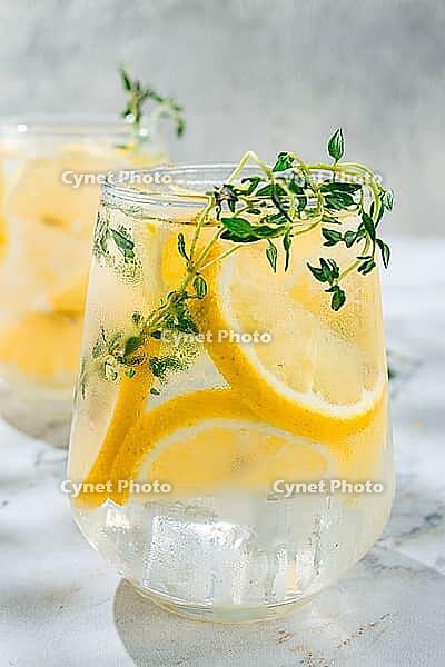 Two clear glasses hold a cold lemon drink with slices of lemon and sprigs of thyme. A half lemon rests nearby on a marble surface. Bright light illuminates the scene [IBR124748980]