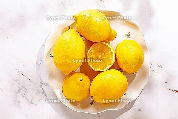 Lemons are arranged on a white decorative plate. One lemon is cut in half, revealing the bright yellow inside. The setting has natural light coming in [IBR124748986]