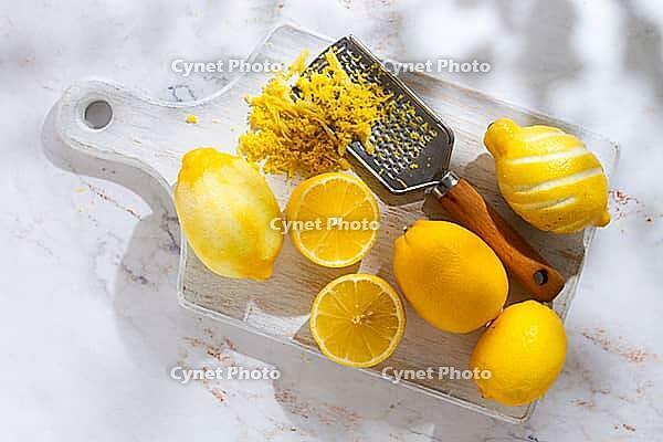 Lemons are placed on a wooden cutting board. Some lemons are whole while others are cut into slices. A zester is used to create lemon zest. The scene shows a bright kitchen [IBR124748995]