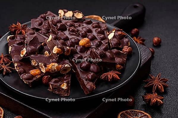 Food, Black bitter chocolate with whole hazelnuts, candied fruits and mint, broken into pieces on a dark concrete background [IBR124773712]