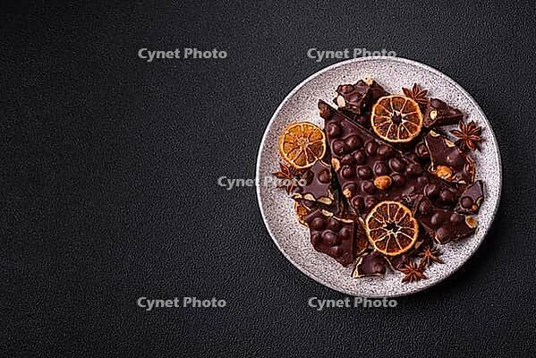 Food, Black bitter chocolate with whole hazelnuts, candied fruits and mint, broken into pieces on a dark concrete background [IBR124773725]