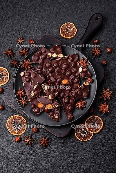 Food, Black bitter chocolate with whole hazelnuts, candied fruits and mint, broken into pieces on a dark concrete background [IBR124773727]