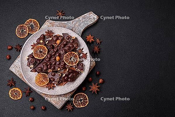 Food, Black bitter chocolate with whole hazelnuts, candied fruits and mint, broken into pieces on a dark concrete background [IBR124773729]
