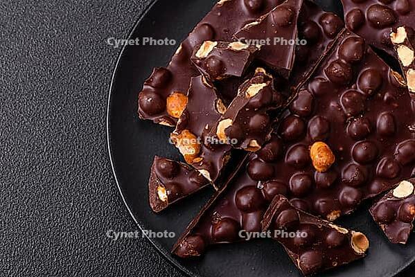 Food, Black bitter chocolate with whole hazelnuts, candied fruits and mint, broken into pieces on a dark concrete background [IBR124773731]