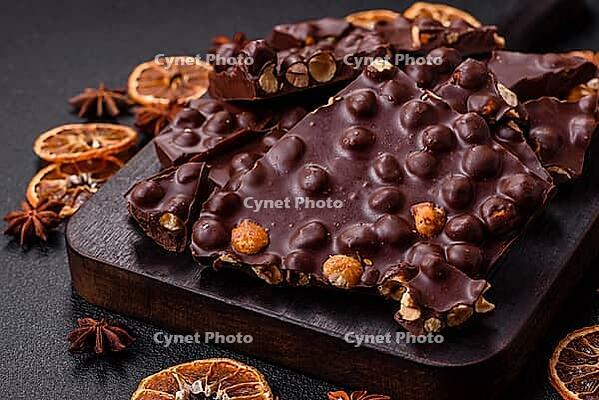 Food, Black bitter chocolate with whole hazelnuts, candied fruits and mint, broken into pieces on a dark concrete background [IBR124773732]