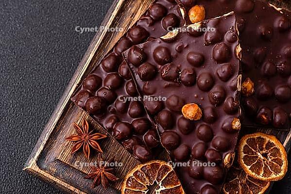 Food, Black bitter chocolate with whole hazelnuts, candied fruits and mint, broken into pieces on a dark concrete background [IBR124773737]