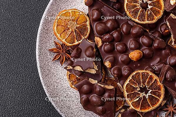 Food, Black bitter chocolate with whole hazelnuts, candied fruits and mint, broken into pieces on a dark concrete background [IBR124773738]