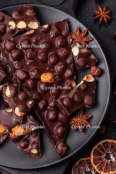 Food, Black bitter chocolate with whole hazelnuts, candied fruits and mint, broken into pieces on a dark concrete background [IBR124773740]