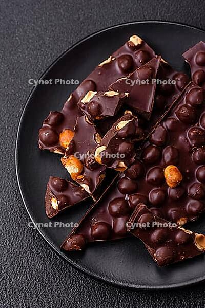 Food, Black bitter chocolate with whole hazelnuts, candied fruits and mint, broken into pieces on a dark concrete background [IBR124773742]