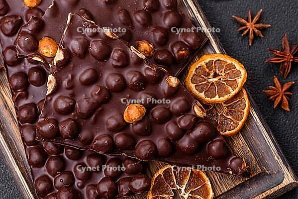 Food, Black bitter chocolate with whole hazelnuts, candied fruits and mint, broken into pieces on a dark concrete background [IBR124773743]