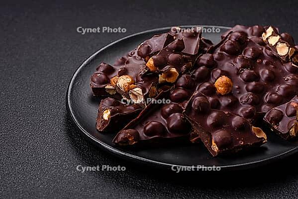 Food, Black bitter chocolate with whole hazelnuts, candied fruits and mint, broken into pieces on a dark concrete background [IBR124773747]