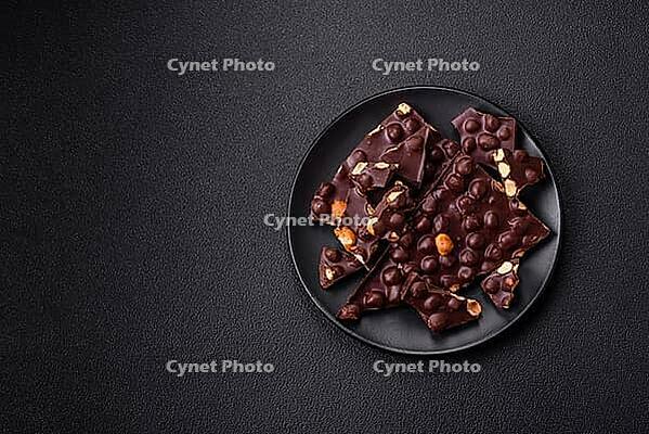Food, Black bitter chocolate with whole hazelnuts, candied fruits and mint, broken into pieces on a dark concrete background [IBR124773748]