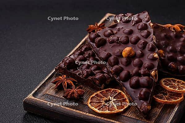 Food, Black bitter chocolate with whole hazelnuts, candied fruits and mint, broken into pieces on a dark concrete background [IBR124773749]