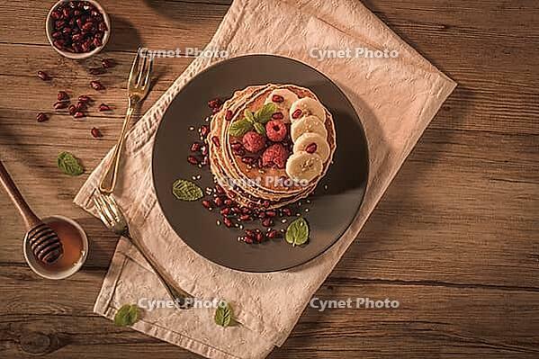 Pancakes with raspberries, banana slices, pomegranate seeds and honey on wooden vintage table [IBR124817897]