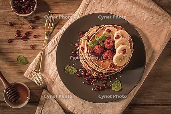 Pancakes with raspberries, banana slices, pomegranate seeds and honey on wooden vintage table [IBR124817898]