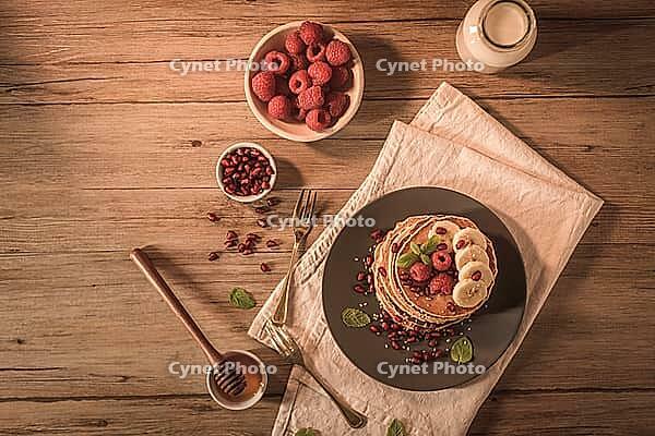 Pancakes with raspberries, banana slices, pomegranate seeds and honey on wooden vintage table [IBR124817901]