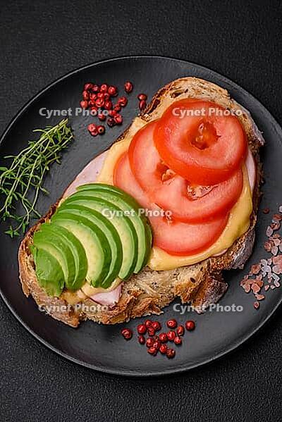 Food, Delicious nutritious grilled toast with ham, cheese, tomatoes and avocado with salt, spices and herbs on a dark concrete background [IBR124842035]