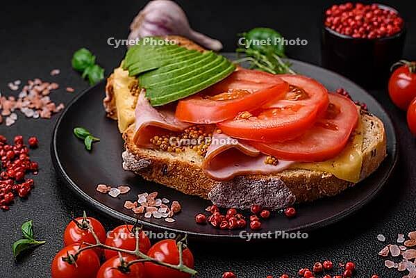 Food, Delicious nutritious grilled toast with ham, cheese, tomatoes and avocado with salt, spices and herbs on a dark concrete background [IBR124842036]