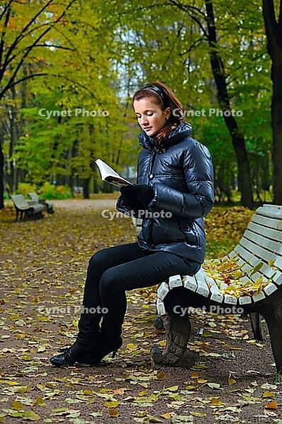 Young woman is sitting on a bench in an autumn park and reading [IBR124906339]
