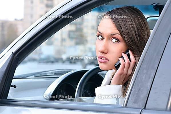 Sensual young woman is looking at camera from a car and talking phone [IBR124906344]