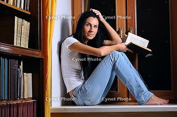 Beautiful woman is sitting on a windowsill and holding a book [IBR124906346]