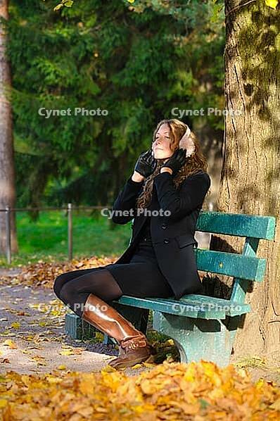 Young woman is sitting on the bench in autumn park and listening music [IBR124906367]