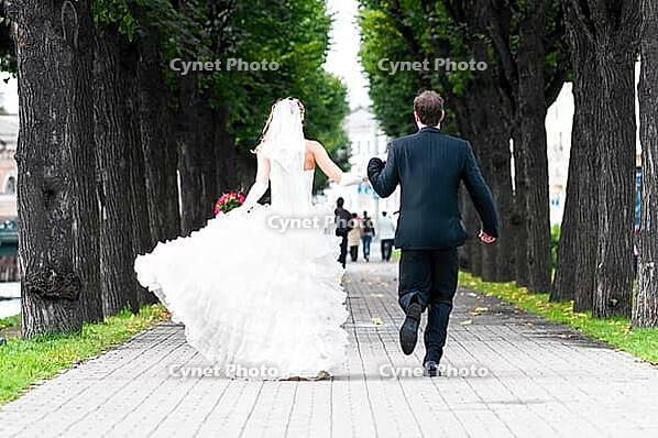 Bride and groom is running with joined hands on city park road [IBR124906369]