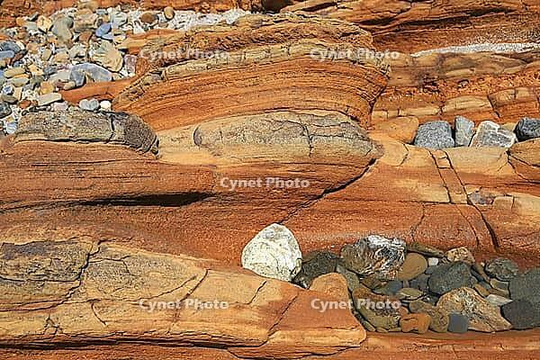 Detailed nature photo of a reddish-brown sandstone formation on a coast. The close-up shows the distinctive texture of horizontal sediment layers formed by centuries of erosion. Embedded in the rocky structure are various smaller, colorful pebbles and tid [IBR124920210]