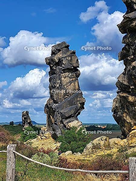 Spectacular view of the historic Devil's Wall in an idyllic heath landscape. The mighty sandstone rock formation fascinates with its rugged structures, earthy tones and a distinctive, free-standing rock needle in the background. A wooden rope fence lines  [IBR124920218]
