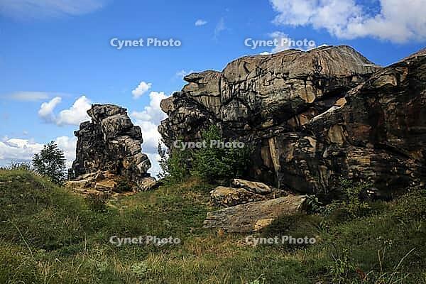 Spectacular view of the historic Devil's Wall in an idyllic heath landscape. The mighty sandstone rock formation fascinates with its rugged structures, earthy tones and a distinctive, free-standing rock needle in the background. A wooden rope fence lines  [IBR124920221]