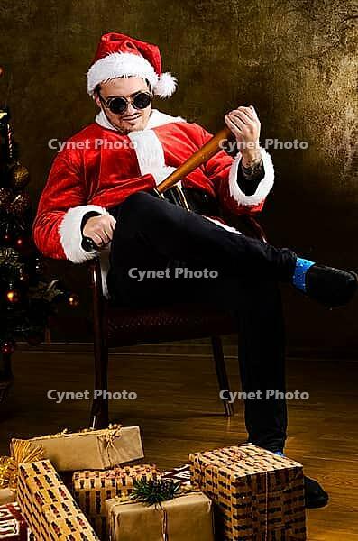 Bad Santa with baseball bat sitting indoors near Christmas or New Year fir tree [IBR124944047]