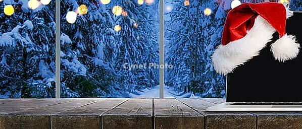 Laptop on table with Santa Claus hat at home with panoramic view through window of snowy trees in winter forest [IBR124944123]