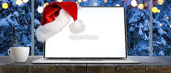 Laptop on table with Santa Claus hat at home with panoramic view through window of snowy trees in winter forest, screen is cut with provided clipping path [IBR124944127]