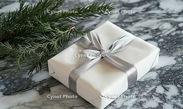Neatly wrapped white gift with a silver ribbon, positioned on a marble table next to a sprig of fir. AI generated [IBR125000271]