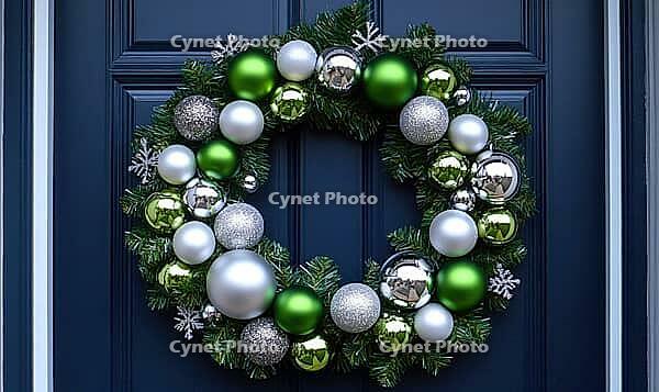 Simple white wreath with silver and green ornaments, hanging on a dark blue door, illuminated by natural daylight. AI generated [IBR125000278]