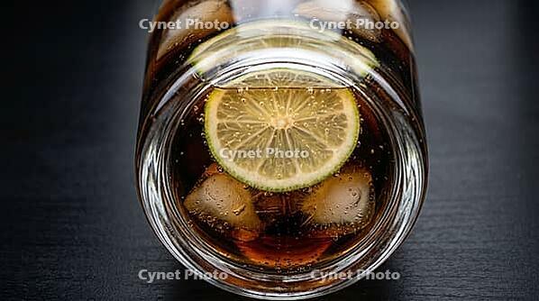 Extreme macro shot looking upward through thick transparent glass base of tall highball glass containing dark rum cola cocktail from below table level, pale green-yellow lime wedge underside visible floating at mid-level with three cloudy ice cubes above  [IBR125000321]