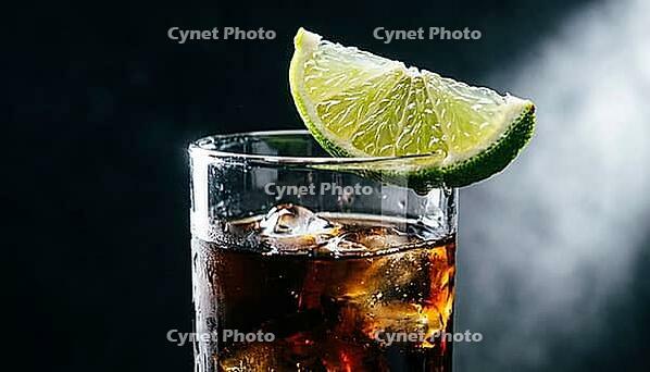 Extreme macro close-up of fresh lime wedge balanced diagonally on rim of tall highball glass filled with near-opaque dark cola rum cocktail, single Dedolight spot from right raking lime skin in full relief revealing every pore and oil cell, glass rim catc [IBR125000322]