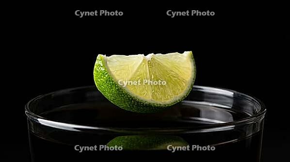 Extreme macro close-up of fresh lime wedge frozen mid-fall between two and four centimetres above perfectly still near-black rum cola cocktail surface inside tall highball glass, lime razor-sharp with every pore visible under raking Dedolight spot, undist [IBR125000324]