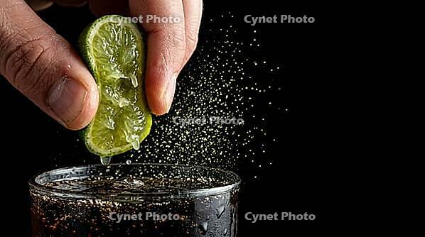 Extreme macro close-up of two fingers squeezing fresh lime wedge over tall highball glass containing dark rum cola cocktail, extraordinary subject is visible golden-green citrus oil micro-mist of ruptured oil cells hanging mid-air between lime and drink c [IBR125000325]